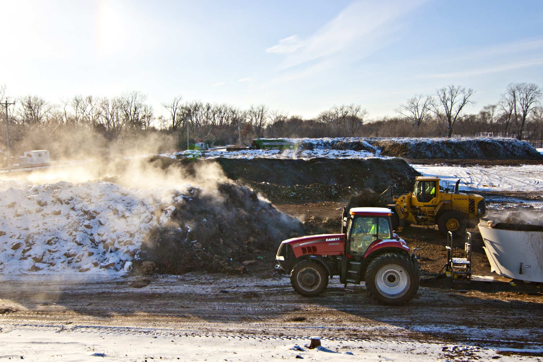 Tractor in a commercial/industrial operation of composting next to a giant pile of steaming compost product